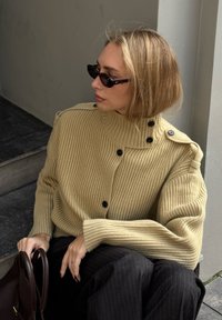 Béžový žebrovaný svetr s vysokým límcem, se dvěma černými knoflíky na ramenou. Modelka má sluneční brýle ve tvaru kočičích očí a drží hnědou tašku.