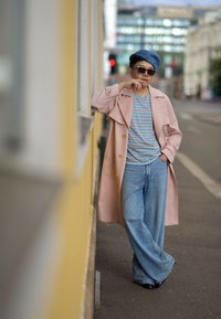 Palton lung roz deschis cu nasturi aurii, cămașă în dungi albastre, beretă albastră deschisă, blugi evazați albaștri și pantofi negri pe un fundal urban.