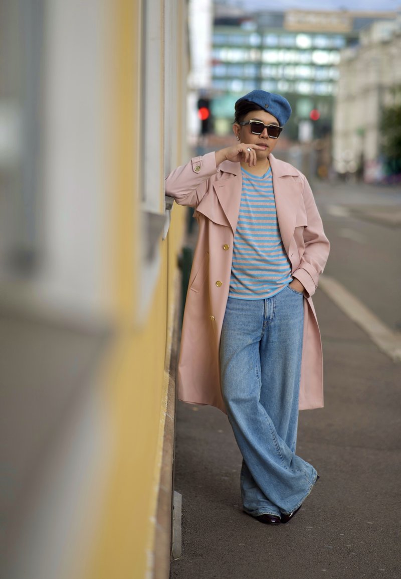 Trench coat rosa chiaro con bottoni dorati, camicia a righe blu, berretto blu chiaro, jeans a gamba larga blu e scarpe scure su uno sfondo urbano.