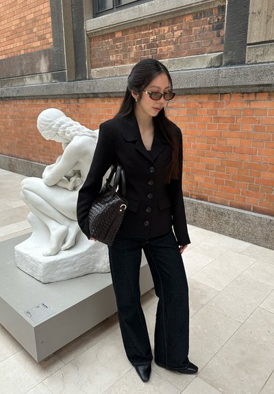 Mujer con blazer negro, gafas de sol y bolso tejido, situada cerca de una escultura de mármol blanco de una figura sentada en una galería con paredes de ladrillo.