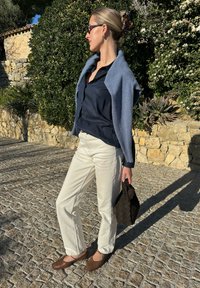 Ανοιχτό μπλε πουλόβερ πάνω από ένα σκούρο ναυτικό μπλουζάκι, λευκά παντελόνια και καφέ μοκασίνια. Κ�ρατά μια υφασμάτινη καφέ τσάντα. Στον ύπαιθρο με πράσινο φόντο.