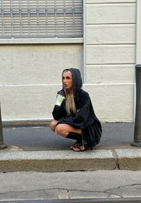 Oversized black hoodie with a hood, worn over dark shorts. Drinking from a black cup with a straw while squatting on a sidewalk, gray background.