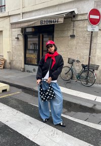 Personne portant une casquette rouge et une écharpe, une veste noire, un jean ample, tenant un sac à main à pois, se tenant dans une rue de la ville près d'un vélo et d'un magasin avec une enseigne "BIONDI".