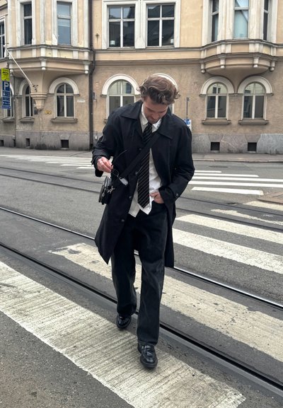 Abrigo negro sobre camisa blanca y corbata de rayas, combinado con jeans oscuros y zapatos negros. Persona camina por un paso de peatones.