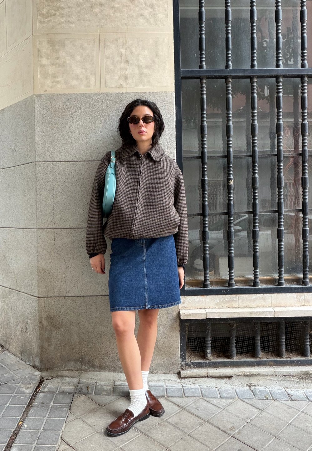 Femme aux cheveux courts et bouclés portant des lunettes de soleil, une veste marron à carreaux, une jupe en denim, des chaussettes blanches et des mocassins marron, debout près d'une fenêtre avec des barreaux.