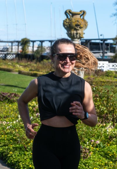 Mujer con gafas de sol y coleta corriendo por un parque, sonriendo, lleva una camiseta negra sin mangas y un reloj deportivo, con vegetación al fondo.