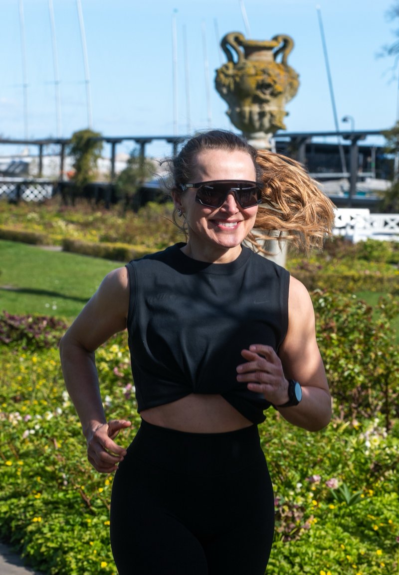 Femme avec des lunettes de soleil et une queue de cheval faisant du jogging dans un parc, souriante, portant un haut noir sans manches et une montre de fitness, verdure en arrière-plan.