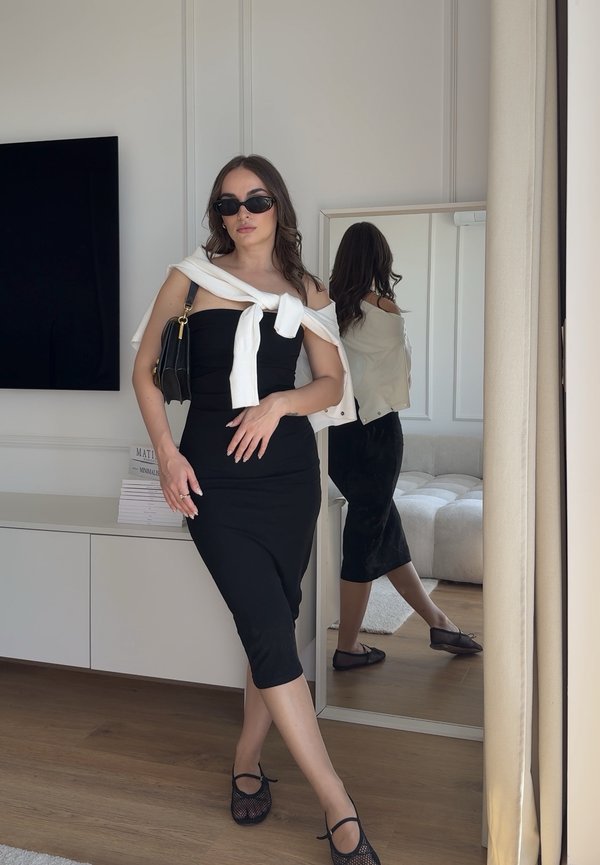 Femeie purtând rochie midi neagră, pulover alb legat peste umeri, poșetă neagră și ochelari de soare, stând în interior lângă o oglindă și un televizor.