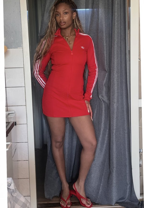 Mujer con el pelo largo trenzado que lleva un vestido rojo de Adidas y sandalias de tacón rojo, de pie en una puerta con cortinas grises.