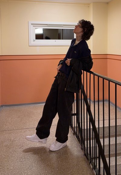 Persona con una camisa polo azul marino y pantalones negros, apoyada en una barandilla negra en un pasillo con paredes amarillas y melocotón. Se ven zapatillas blancas.