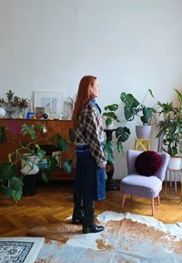 Γυναίκα με κόκκινα μαλλιά φορούσα καρό σακάκι και μαύρα μπότες στέκεται σε δωμάτιο γεμάτο φυτά με ξύλινο πάτωμα και μοβ καρέκλα με στρογγυλό μαξιλάρι.