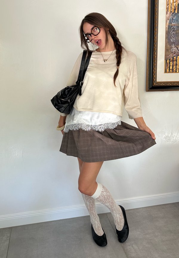 Joven mujer con gafas que lleva una camiseta crema, falda con encaje, calcetines hasta la rodilla estampados, zapatos negros y lleva un bolso negro de hombro posando en el interior.