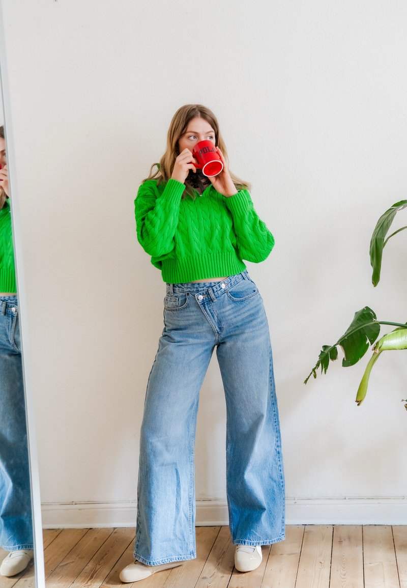 Joven mujer con suéter verde brillante y vaqueros de pierna ancha bebe de una taza roja, de pie cerca de un gran espejo y una planta de interior.