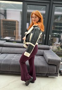 Femme aux cheveux roux portant une veste en peau lainée noire et blanche et un pantalon en tricot violet, posant à l'intérieur près d'un canapé gris et de grandes fenêtres.