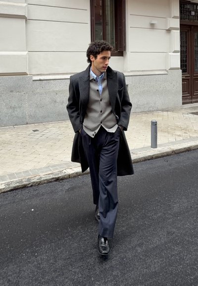 Hombre caminando por la calle de la ciudad con un abrigo negro, chaleco gris, camisa azul claro, pantalones azul marino y zapatos negros con las manos en los bolsillos.