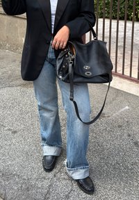 Osoba nosząca podwinięte jasnoniebieskie dżinsy, czarną marynarkę i czarne mokasyny, trzymająca dużą czarną skórzaną torebkę z metalowymi zapięciami.