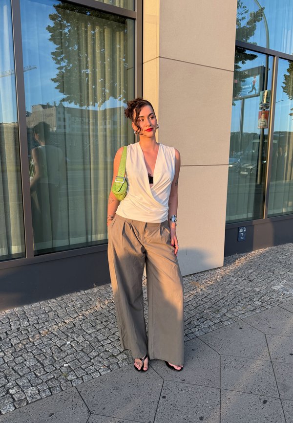 Jeune femme en haut blanc sans manches, pantalon large beige et sandales, appuyée contre un mur de bâtiment avec un sac à bandoulière vert.