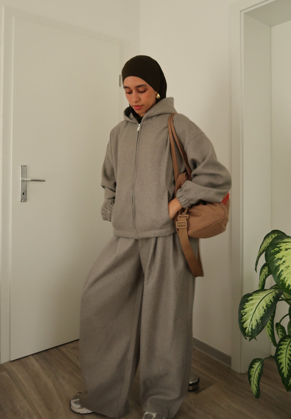 Sweat à capuche gris surdimensionné avec fermeture éclair et poignets côtelés, accompagné d'un pantalon de survêtement gris à jambes larges. Sac bandoulière marron et baskets complètent le look.