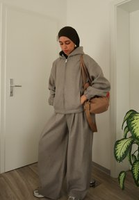 Sweat à capuche gris surdimensionné avec fermeture éclair et poignets côtelés, accompagné d'un pantalon de survêtement gris à jambes larges. Sac bandoulière marron et baskets complètent le look.