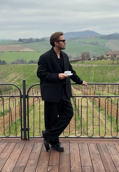 Traje negro a medida, camisa negra y gafas de sol, llevados por un hombre que sostiene una taza blanca. El fondo presenta viñedos verdes y colinas onduladas.