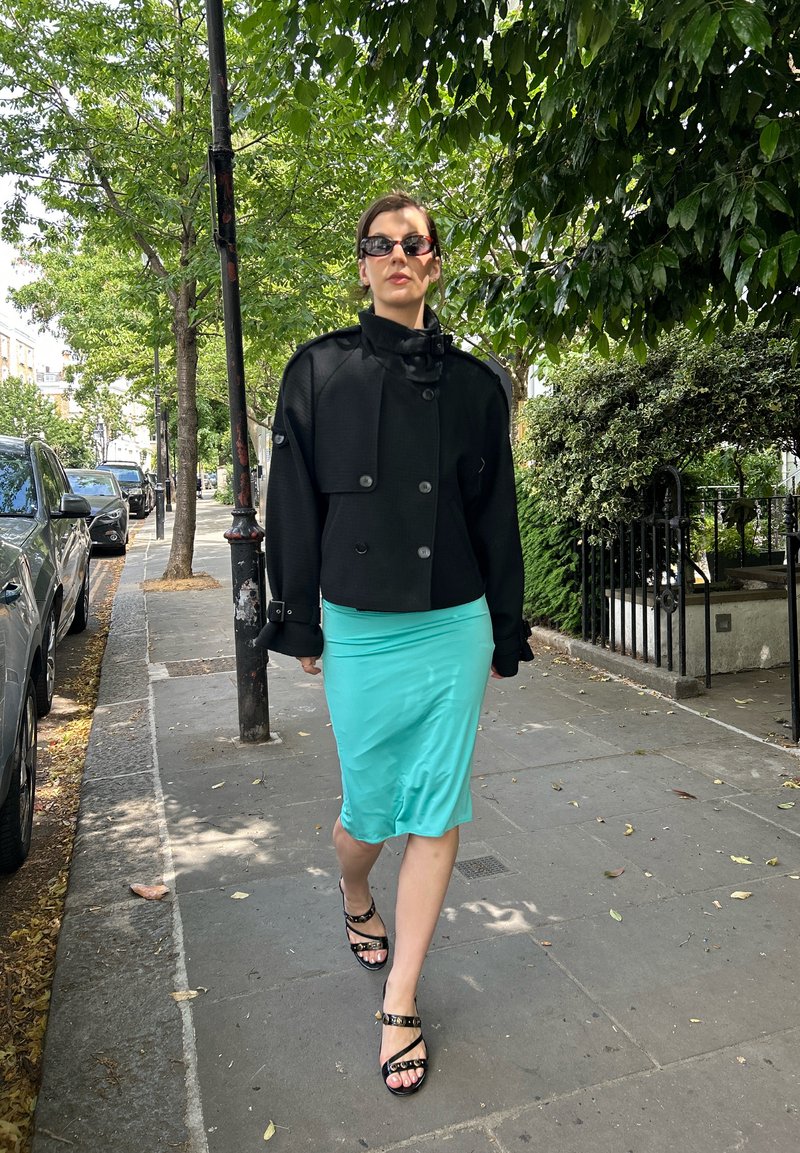 Femme portant une veste noire, une jupe turquoise et des sandales noires marchant sur un trottoir bordé d’arbres en ville pendant la journée.