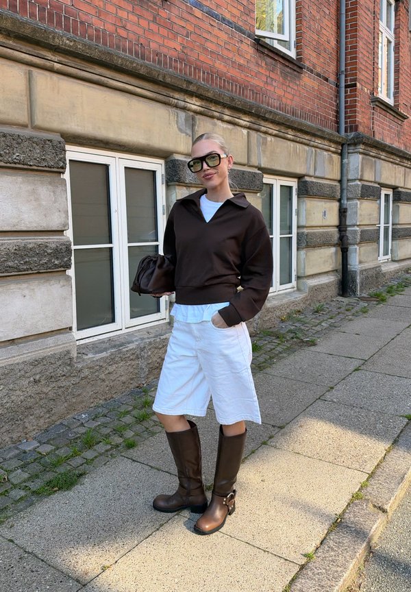 Una mujer con gafas de sol, un jersey oscuro, pantalones cortos blancos y botas altas marrones posa en una acera de la ciudad junto a un edificio de ladrillo.