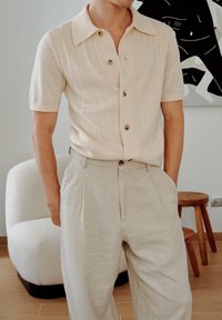 Camisa beige com riscas, de manga curta, colarinho pontudo e fecho de botão, combinada com calças de linho cinza claro. Textura suave, corte descontraído.