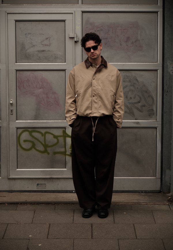 Homme portant une veste beige, un pantalon large noir et des lunettes de soleil, debout devant une porte métallique recouverte de graffiti sur un trottoir en ville.