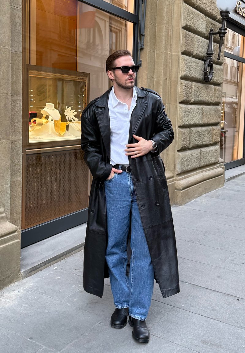 Homme portant un trench-coat en cuir noir, une chemise blanche, un jean bleu et des bottes noires se tient sur le trottoir de la ville près d'une vitrine avec présentation de bijoux.