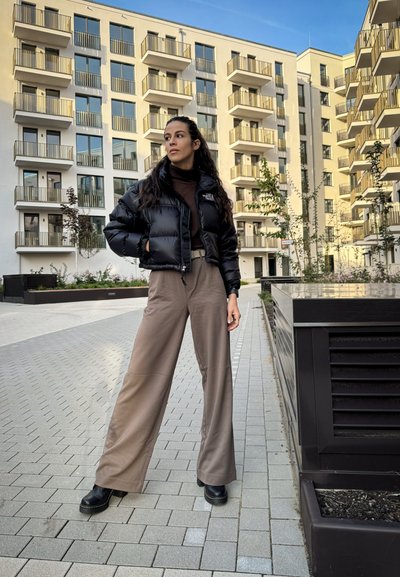 Chaqueta de plumas negra, jersey de cuello alto marrón, pantalones anchos beige y botas de plataforma negras. Entorno urbano moderno con balcones de fondo.