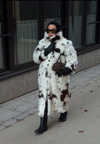 Hosszú műszőrme kabát fehér és barna foltokkal, fekete kesztyűk, keskeny táska és fekete gumicsizma, visszafogott városi háttér előtt.