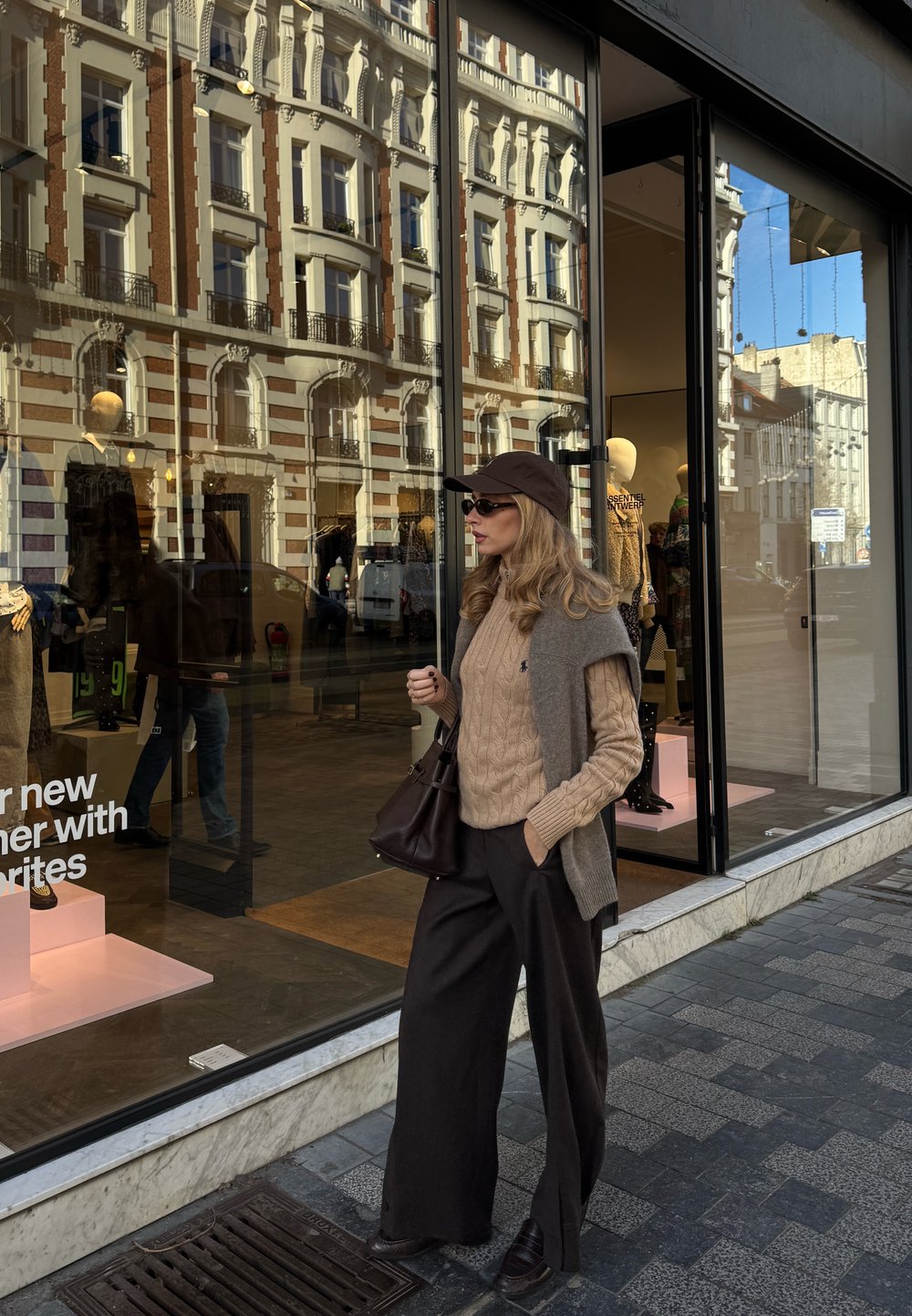 Strikket beige genser, grå vest og vidbukser i svart. Mørk brun håndveske, matchende caps og solbriller. Refleksjon av butikkskap synlig.