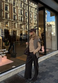 Strikket beige genser, grå vest og vidbukser i svart. Mørk brun håndveske, matchende caps og solbriller. Refleksjon av butikkskap synlig.