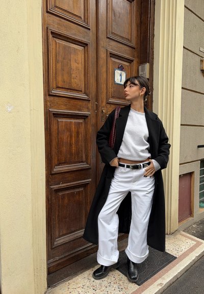 Abrigo largo negro, suéter gris, pantalones anchos blancos, cinturón negro y botines negros. Puerta de madera con detalles en panel en el fondo.