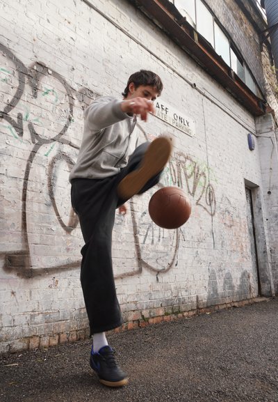 Persona con sudadera gris y pantalones negros pateando una pelota marrón contra una pared de ladrillos blancos cubierta de graffiti en un callejón.