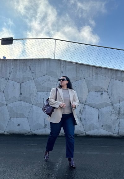 Blazer beige sobre una blusa clara, jeans oscuros y botines oscuros. Bolso marrón. El fondo presenta una pared de concreto texturizada y cielo azul.