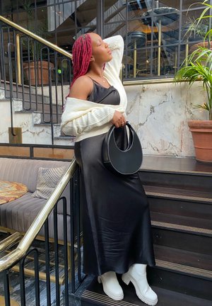 Vestido de satén negro con una silueta fluida, cárdigan de punto blanco, bolso negro y botas de plataforma blancas. Lleva el cabello trenzado en rosa.