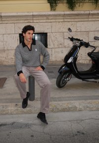 Chandail à motifs gris et noir, pantalon en velours côtelé beige ample, chaussures noires, assis sur un poteau en béton, avec une trottinette à proximité.