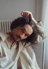 Persoon met krullend haar, gekleed in een licht beige shirt en een zilveren ketting, zittend tegen een zacht textuur achtergrond met een radiator.