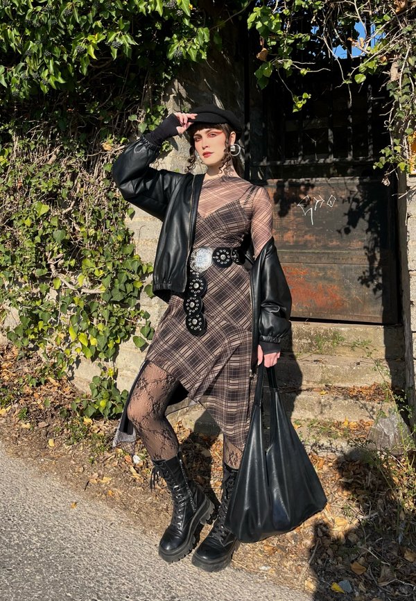 Persoană îmbrăcată într-o rochie asimetrică în carouri, jachetă neagră de piele, beretă, dresuri din dantelă, bocanci de luptă și curea mare, stând lângă trepte de piatră cu vegetație.