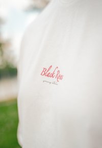 Vit bomull T-shirt med en liten röd broderad logotyp "Black Roses" och grå text nedanför, mot en suddig utomhusbakgrund.