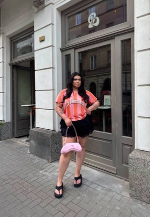 Camisa Adidas a rayas rosas y blancas, falda negra con capas, chanclas de plataforma negras y un bolso rosa claro, de pie en una acera pavimentada.