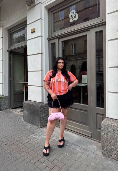 Camisa Adidas a rayas rosas y blancas, falda negra con capas, chanclas de plataforma negras y un bolso rosa claro, de pie en una acera pavimentada.