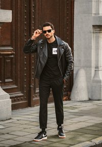 Fekete bőr motoros dzseki egy fekete póló felett, fehér logóval, fekete nadrág és fekete sportcipő, fehér és narancssárga díszítéssel.