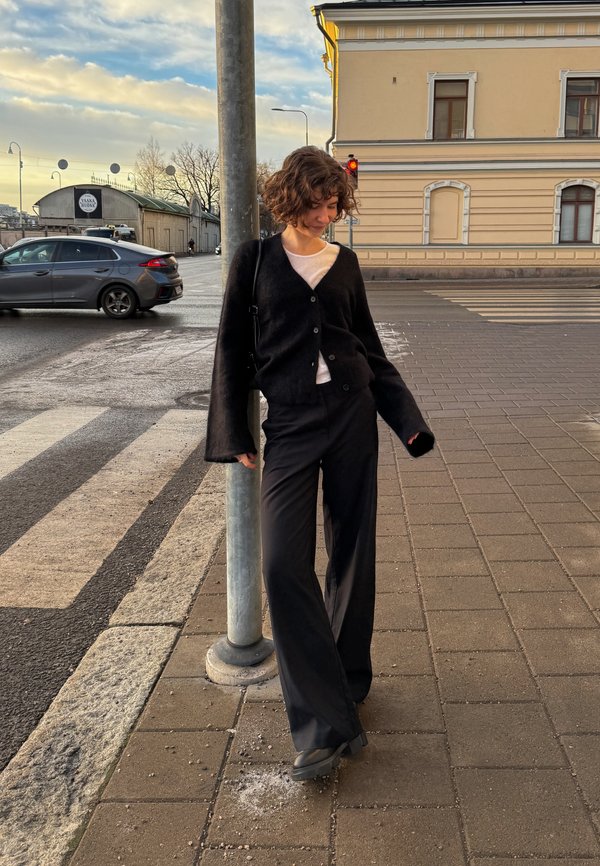 Black cardigan with long sleeves, white t-shirt, and black wide-leg trousers. Standing by a pole on a paved street. Soft textures and neutral colours.