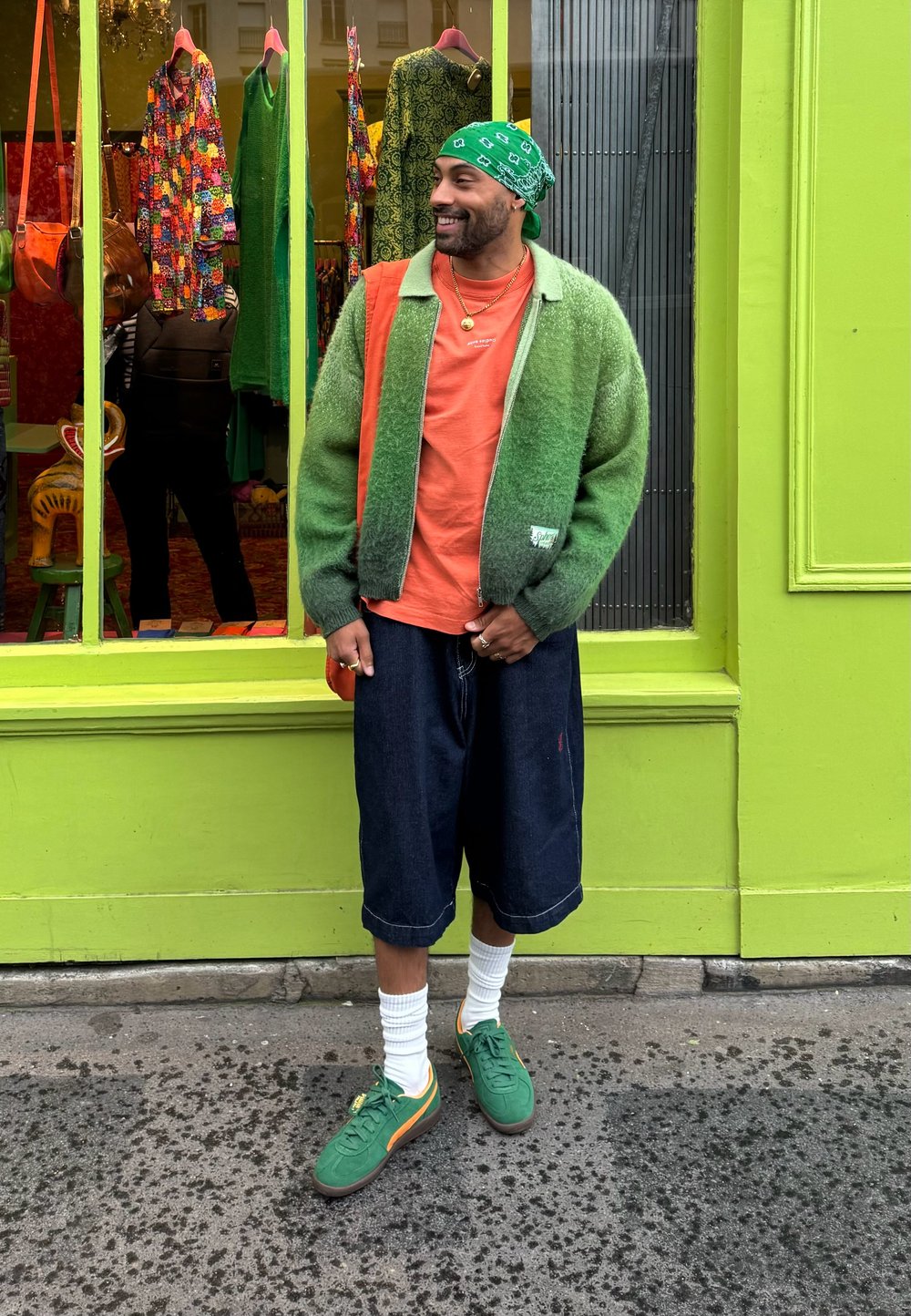 Πράσινο gradient φλουώδες μπουφάν πάνω από πορτοκαλί μπλούζα, σκούρες τζιν βερμούδες, λευκές κάλτσες και πράσινα αθλητικά παπούτσια; πολύχρωμα μοτίβα ρούχων στο παρασκήνιο.