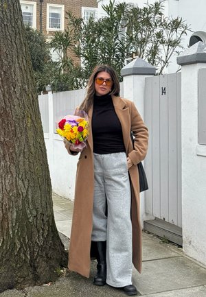 Mujer con abrigo camel, cuello alto negro, pantalones de chándal grises y gafas de sol sosteniendo un ramo de flores colorido junto a una puerta blanca con el número 14.