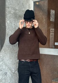 Suéter marrón de punto con un corte relajado, usado sobre pantalones negros, complementado con un gorro negro de punto y gafas, posando en un entorno con textura.