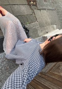 Mujer con cabello castaño y gafas de sol, vistiendo una camisa de cuadros y una falda de lentejuelas transparente, sentada en escalones de piedra al aire libre junto a una puerta de madera.