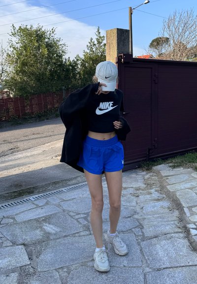 Sudadera cortada negra con el logo blanco de Nike, pantalones cortos deportivos azules, gorra de béisbol blanca y zapatillas blancas sobre un pavimento de piedra.